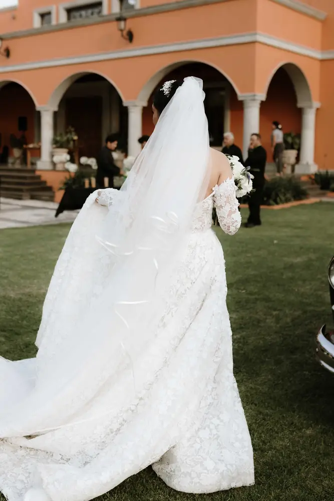 Novia caminando hacia los arcos de la hacienda con el velo extendido sobre el pasto